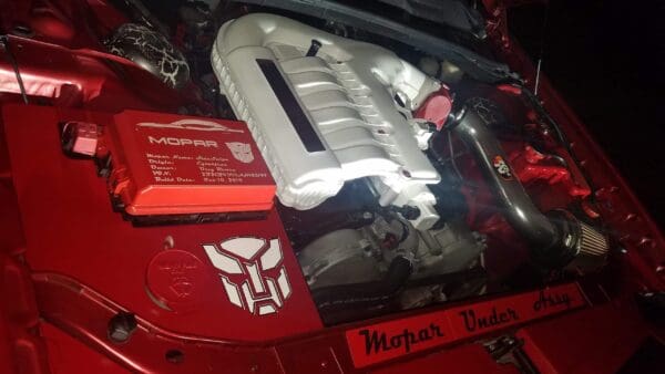 The engine of a red car with the hood up, featuring Mopar branding and a Transformers logo.