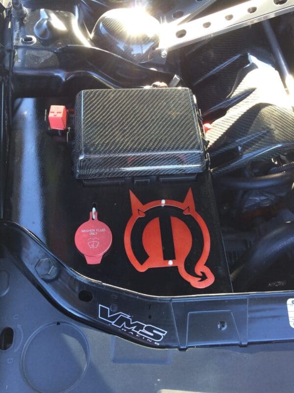 Close-up of a car engine featuring a carbon fiber cover, red "Washer Fluid Only" cap, and a red devil logo. VMS Racing branding visible.