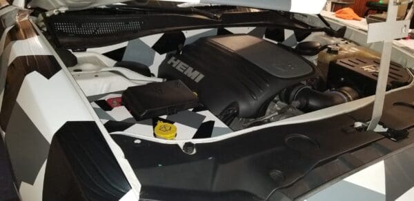 Car engine with hood up, featuring a HEMI engine cover and geometric black and white pattern on the fender.