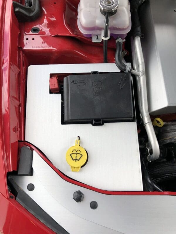The engine compartment of a red and white car, featuring a yellow windshield washer fluid cap and a black fuse box.