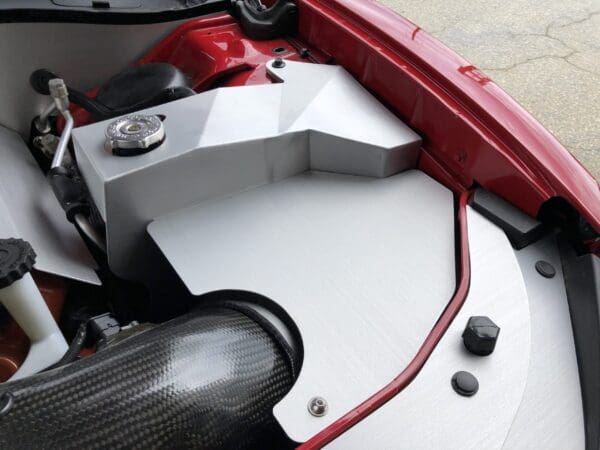 Close-up of a car's engine compartment, featuring a sleek, silver cover and carbon fiber components against a red frame.