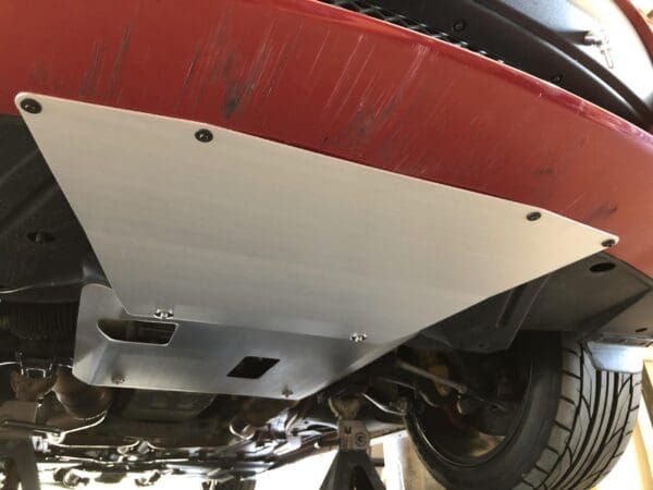 Silver skid plate installed beneath a red car, showing the tire and suspension components.