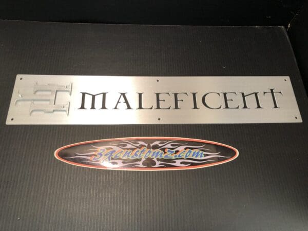 Silver "Maleficent" informational sign above a "3customz.com" sticker on textured surface.