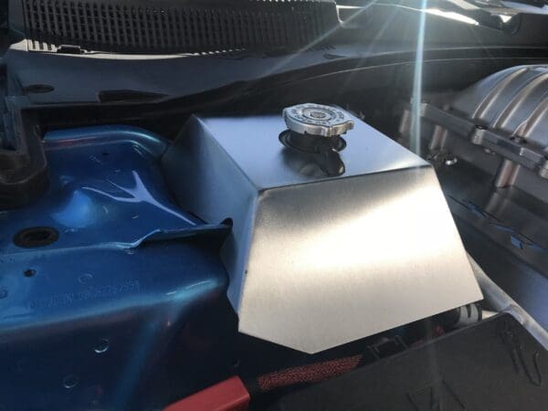 Close-up of a car engine showing a metal cover with a cap, adjacent to a blue component and part of the intake manifold.