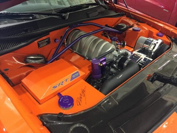 The engine compartment of an orange sports car features an SRT engine with purple accents and a carbon fiber cover.