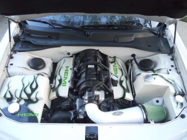 The engine compartment of a white car is shown, featuring a HEMI engine with custom-painted covers and green flame designs.