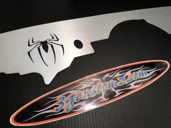 ```plaintext
Close-up of a skateboard with a spider design and a 3gcustomz.com sticker on a black surface.
```
