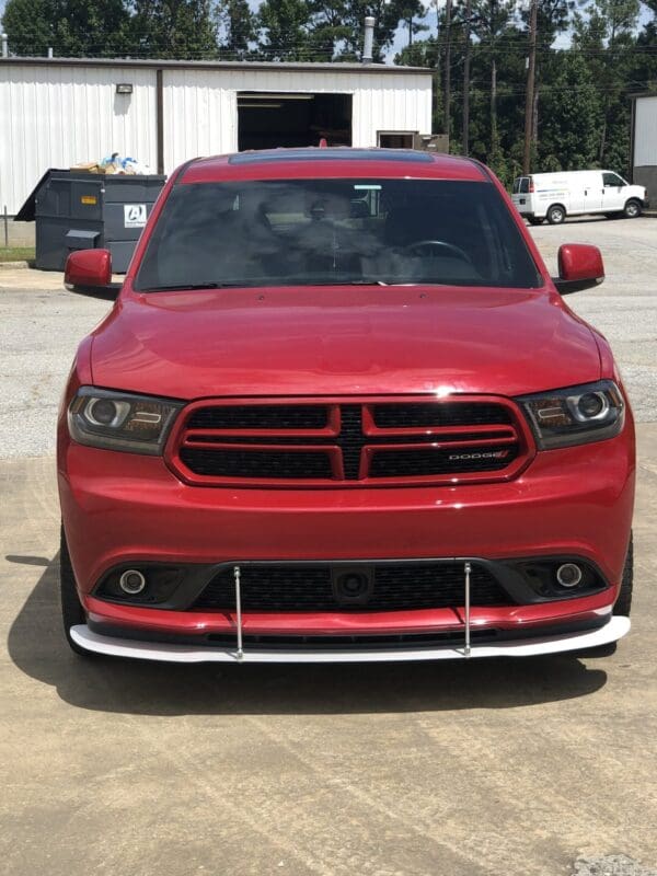 A red Dodge Charger is parked in a lot, with industrial buildings and vehicles in the background.