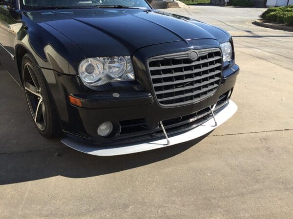 A black car with a custom front splitter is parked in a driveway on a sunny day.