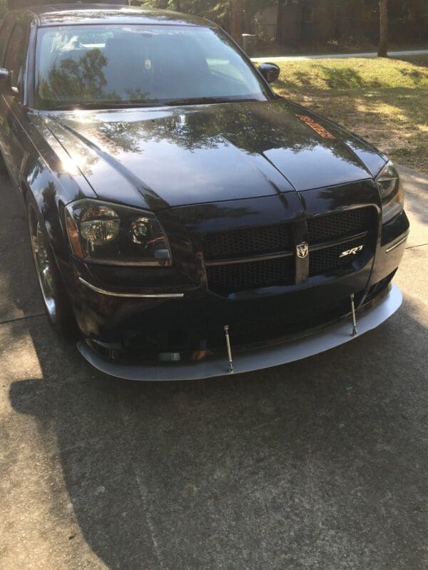 A black Dodge Magnum SRT parked on a driveway, reflecting sunlight and surrounded by greenery.
