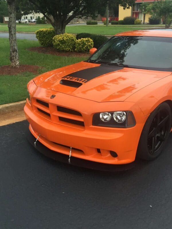 An orange Dodge Charger with a HEMI hood parked in a lot, surrounded by greenery and buildings in the background.
