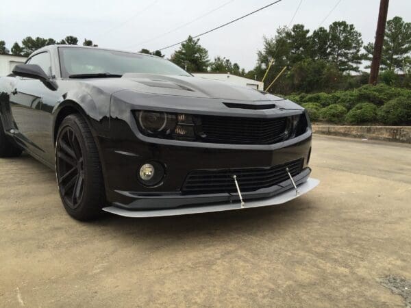 A sleek black sports car with a modified front splitter parked in a lot, surrounded by greenery and a cloudy sky.