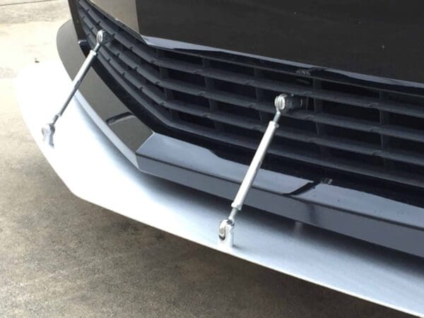Close-up of a car's front bumper with a black grille and two stabilizing rods attached to a white splitter.