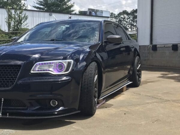 A black car with purple headlights is parked in front of a building, featuring tinted windows and custom wheels.