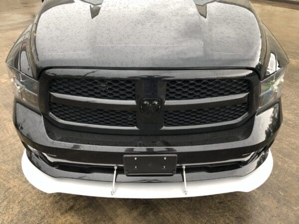 The front of a gray truck with a black grille, parked in a lot, showing a license plate holder and white trim below.