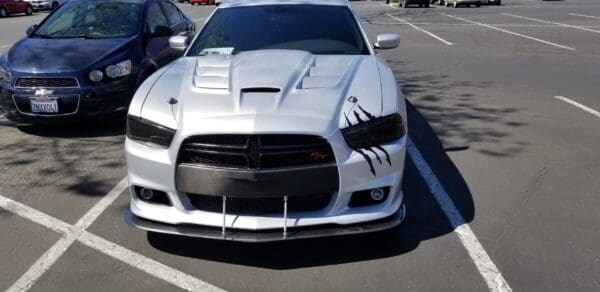 Two cars parked side by side in a parking lot; a blue Chevrolet and a white Dodge Charger with distinctive hood details.
