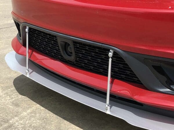 Close-up of a red car's front bumper featuring a black grille and a silver splitter with support rods.