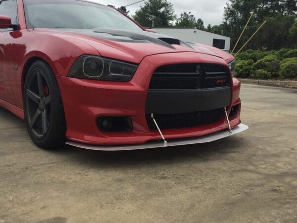 A red Dodge Charger SRT with a custom front splitter is parked in a lot, surrounded by greenery and a building in the background.