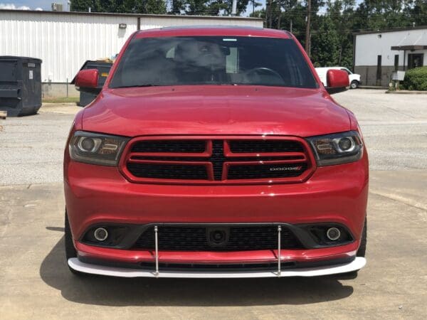 A red Dodge Charger is parked in a parking lot, with industrial buildings and dumpsters in the background.