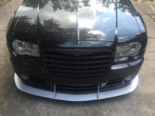 A black car with a reflective hood and a front splitter, parked on a concrete surface.