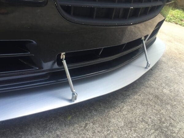 The front bumper of a black sports car with a silver splitter and supporting rods.
