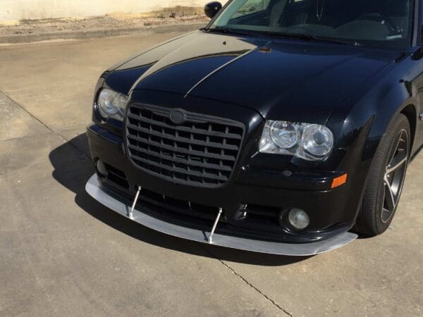 A black car with a custom front splitter parked in a lot, highlighting its grille and headlights.