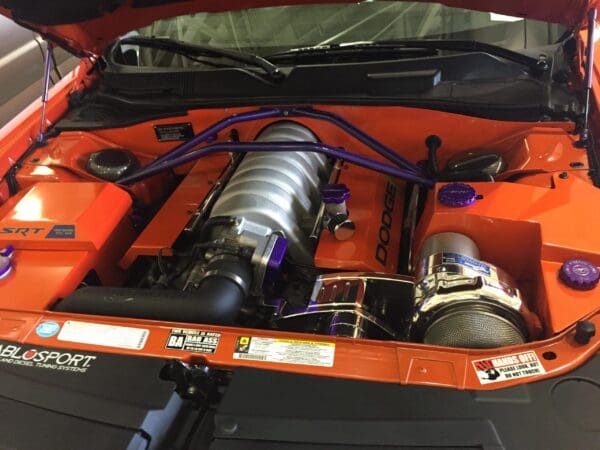 The engine compartment of an orange Dodge car with its hood open, showcasing a powerful setup with purple accents.