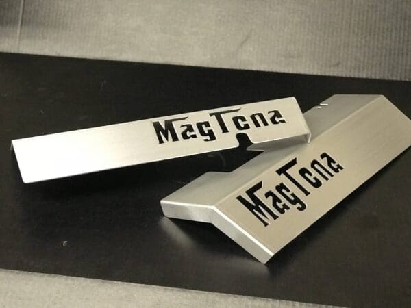 A pair of silver MagTena magnets on a black table, displaying the logo in bold cut-out letters.
