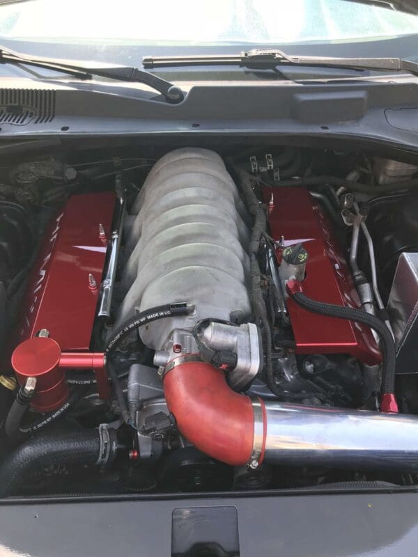 The open hood reveals a car's engine compartment, featuring a prominent intake manifold and red valve covers.
