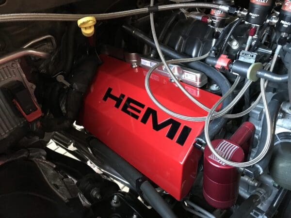 Close-up of a car engine featuring a red cover with "HEMI" in bold black letters, surrounded by metal hoses.