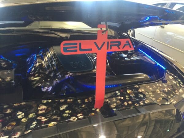 A black car with the hood open and a hood prop that reads ELVIRA