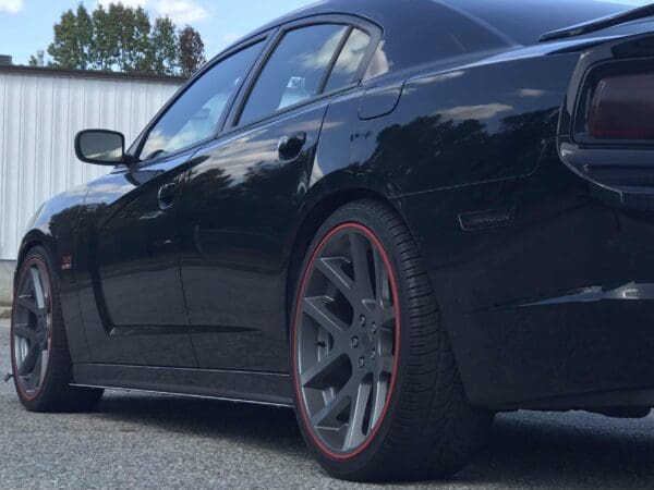 A black Dodge Charger parked in a lot, featuring red-trimmed gray rims and a spoiler, with a white building in the background.