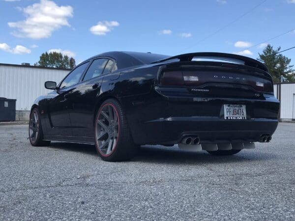 A black Dodge Charger parked in a lot, featuring large gray wheels with red rims, against a backdrop of a white building and blue sky.