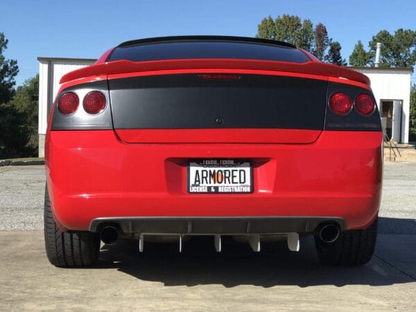 A red sports car with a black rear panel and custom license plate reading "ARMORED," parked in a driveway.