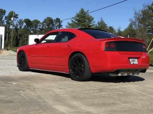 A red sports car with a license plate reading "ARMORED" parked in a lot, surrounded by trees and buildings.