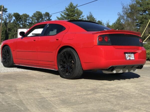 A red sports car with black wheels parked in a lot, surrounded by trees, under a clear blue sky.