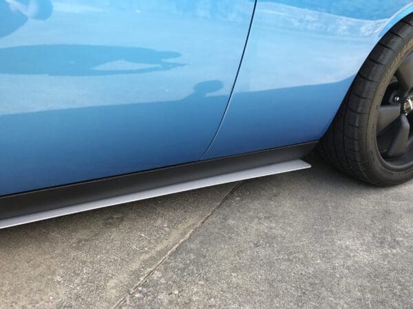 A close-up of a blue car's side, parked on a road, showing the wheel and car body reflection.