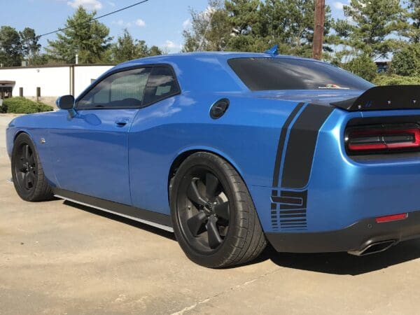A blue sports car with black accents parked in a lot, surrounded by trees and buildings in the background.