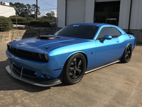A blue Dodge Challenger parked in front of a building with a roll-up garage door and greenery in the background.