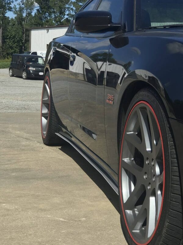 A black sports car with red-trimmed rims parked in a lot, with another vehicle and building in the background.