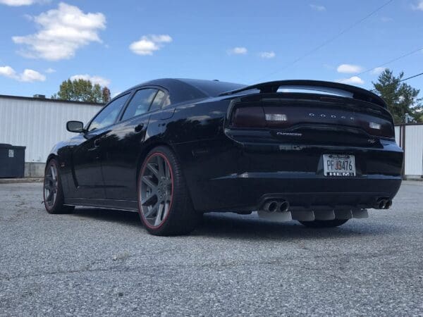 A black Dodge Charger R/T with license plate PF 33476 is parked in a lot, featuring custom rims and dual exhausts.