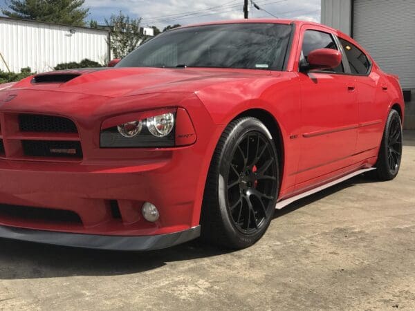 A red Dodge Charger SRT8 parked on a concrete surface in front of a white building with greenery and trees nearby.