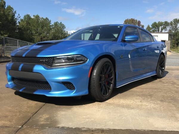 A blue Dodge Charger with black racing stripes is parked in a driveway on a sunny day.