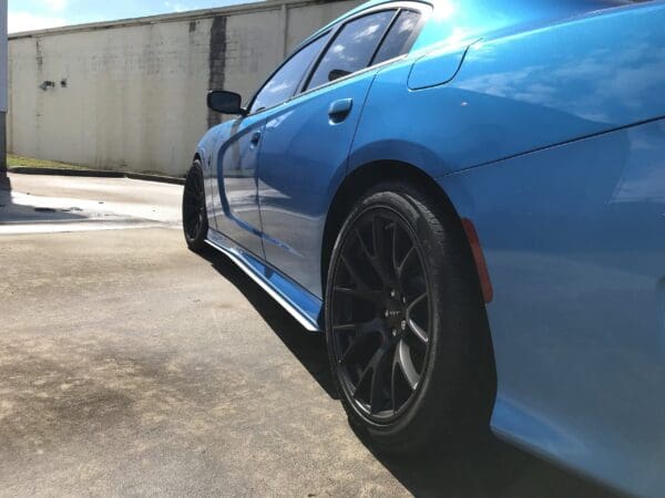 A blue sports car with black rims parked in a sunlit parking lot beside a white brick wall.