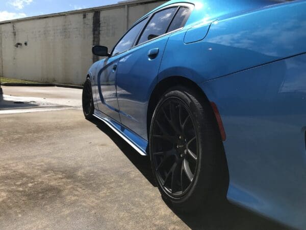 A blue sports car with black wheels parked in a parking lot, next to a white building under a clear sky.