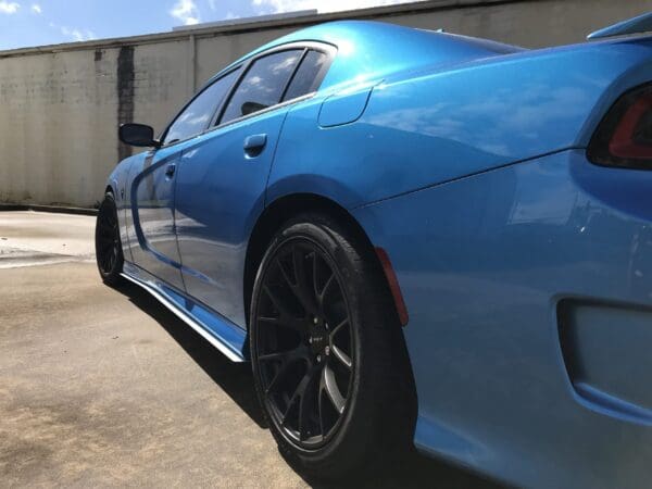 A blue sports car with black rims parked in a parking lot, viewed from a low angle on a sunny day.