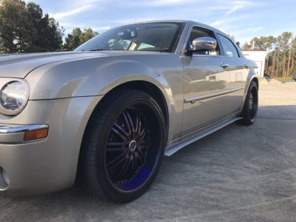 A silver car with custom black wheels parked in a lot, surrounded by trees under a clear sky.