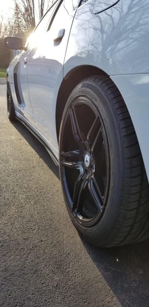 Close-up of a car tire with black alloy wheel, reflecting sunlight on a paved surface.