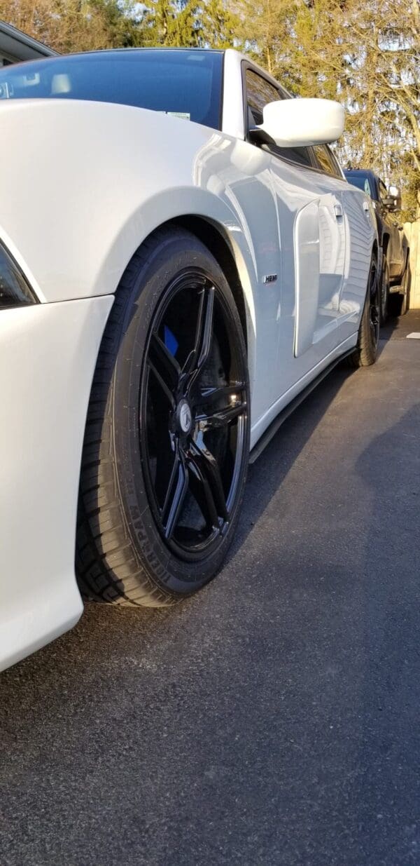 A white sports car with black rims is parked on the side of the road, surrounded by trees.