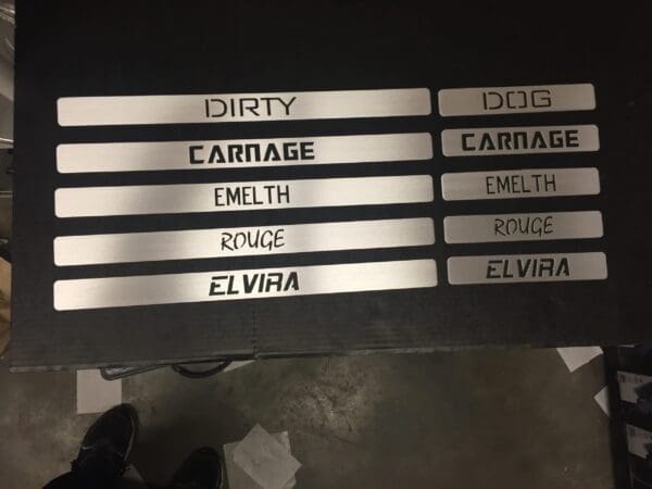 Metal strips with cutout words: DIRTY, DOG, CARNAGE, EMELTH, ROUGE, ELVIRA, displayed on a dark surface.